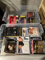 Two clear plastic bins filled with a variety of CDs and DVDs, showing front covers of some music albums and instructional materials.