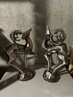 Pair of shiny metal abstract deer figurines with curling antlers and geometric bases placed on a concrete background surface.