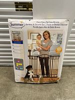 Photo of box front showing woman and baby standing behind bronze metal gate with dog on floor in front. Indicates decorative extra tall safety gate for pets and children by Summer brand.