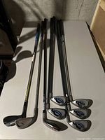 Seven golf clubs displayed horizontally on a table, including irons and a wood, showing the grips and shafts.