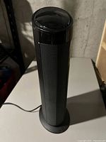 Front view of a slim, black, tower-shaped heater with mesh front and circular base, showing the product in general.