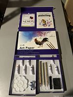 Photo showing open painting set with art paper, instruction booklet, paint tubes, paintbrushes, palette, and ink stone.