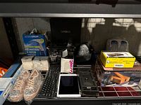 Miscellaneous lot displayed on metal shelf including various boxed items, pair of studded shoes, keyboard, and accessories.