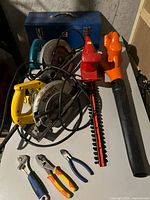 Full view of mixed tools on white surface under dim light, showing two circular saws, hedge trimmer, leaf blower, hand tools, and tool box.