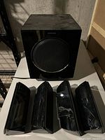 Four black glossy speaker stands or mounting brackets arranged in front of the subwoofer speaker, showing accessories included in the lot.