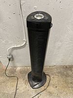 Tower fan plugged in against concrete wall on floor, showing full front view and power cord connected to outlet.