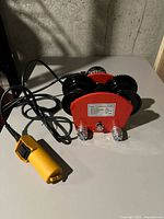 Red single track electric trolley with four black wheels and yellow remote control on a concrete floor.