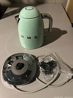 Smeg kettle body, base with power cord, and replacement parts in plastic bag laid out on table for inspection
