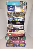 Stack of ten boxed puzzles and WE Games roll-up mat showing titles and piece counts