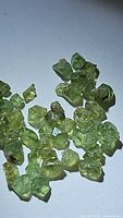Close-up image of roughly 30 small light green demantoid garnet rough crystals scattered on a white background.