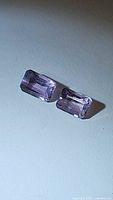 Pair of rectangular emerald cut pink purple amethyst gemstones on a light surface.