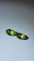 Pair of natural apple green peridot gemstones lying on a flat white surface, showing oval cut and bright green color.