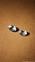 Two natural pear cut aquamarine gemstones on a brown textured surface, showing facets and light reflection.