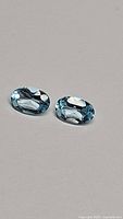 Pair of natural sky blue oval cut topaz gemstones on a plain background showing facets and color.