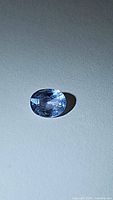 Oval cut denim blue sapphire gemstone lying flat on a light surface under neutral light, showing the top facet and color.