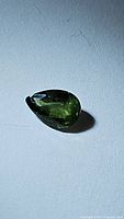 Close-up photo of a 0.83 carat pear-shaped green sapphire on a light surface, showing its rich green color and facets.