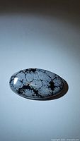 Top view photo of the polished oval snowflake obsidian cabochon with distinct white snowflake patterns on black background.
