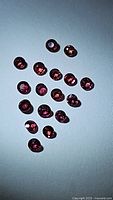 Top view showing 18 small round faceted rubies of deep red color with pink undertones, laid out on a flat surface.