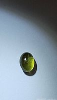 Oval cabochon deep green catseye peridot gemstone lying on a white surface under lighting, highlighting the catseye effect shown as a line across the gem.