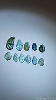 Photo showing 11 leaf-shaped Paraiba blue tourmaline gemstones arranged in two rows with a range of blue and green hues under bright light.