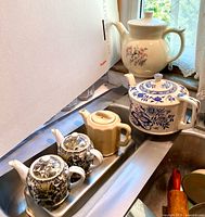 Five teapots in sink: cream floral, blue/white, cream hexagonal, two small black/white/yellow.