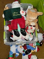 Top-down view of plastic bin filled with various Christmas-themed plush toys including Santa Claus holding a 'Merry Christmas' bag, snowmen with accessories, and a reindeer plush.