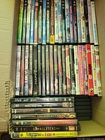 Top-down photo showing a packed box of approx 70-80 pre-owned DVDs with intact cases, mixed genres, stacked in rows. Titles visible range from The Haunting, Happening, to 17 Again and various animation movies.