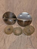 Two round wall-mounted metal ashtrays and three vintage glass ashtrays displayed on a fabric background. Metal ashtrays show minor dents and scratches. Glass ashtrays show wear, discoloration, and staining.