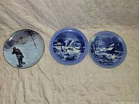 Three decorative blue and white plates with winter scenes laid out on a light fabric background.