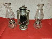 Set of three vintage oil/kerosene lanterns on red surface; two clear glass lanterns with decorative bases flank a metal lantern with protective frame.