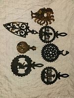 Full view of 7 vintage cast iron trivets arranged on quilted surface showing various folk art designs, rust, and patina.
