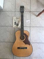 Full view of Marquis by Harmony acoustic guitar laid flat on tile floor with instruction booklet beside it.