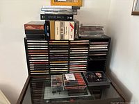 Photo shows black shelving unit loaded with multiple rows of CDs in jewel cases, a few VHS tapes and cassette boxes stacked on top, and some CDs placed on a glass table surface in front.