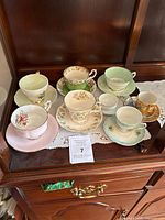 Photo of 8 vintage and rare bone china teacups and saucers displayed on a wooden shelf with crocheted doily under them. Assorted floral patterns, pastel colors, and gold trimming visible.