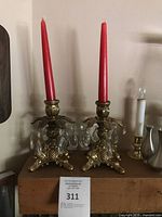 Pair of ornate vintage brass candle sticks with hanging clear crystal prisms and red taper candles on top, displayed on wooden surface.