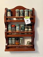 Front view of vintage wooden spice rack mounted on wall with 14 glass spice jars arranged on three shelves. The jars have assorted lids and labels. A tag is attached to the middle shelf rail.