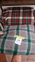 Two folded wool blankets; one red tartan with fringes on top shelf and one grey-green plaid underneath flat on wooden surface with tag #150.