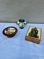 Three music boxes grouped: round Reuge, ceramic Ardleigh Elliott, rectangular Hummel
