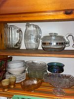 Shelf with two pitchers, glass coffee pot, stacks of plates and bowls, carnival glass dishes and egg cups