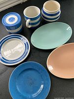 All seven ceramic pieces grouped: striped canister, mug, rounded jar, striped-rim plate, solid blue plate, mint green oval plate, peach oval plate