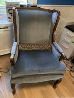 Front view of antique wingback chair upholstered in blue velvet with wooden frame edges and curved cabriole legs, including small floral tapestry pillow at base of backrest.