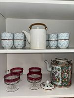 Shelf with ornate Asian ceramic teapot with floral bird design and lid, accompanied by six red Thumbprint dessert glasses on clear stems.