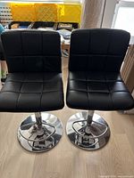 Front view of a pair of black faux leather bar stools with chrome round bases and footrests.