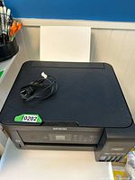 Front view of Epson ET-2750 printer showing control panel, paper tray with paper loaded, and power cord placed on top.