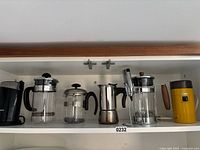 Photo of three sizes of French press coffee makers, stainless steel milk frother, black Krups coffee grinder, and yellow Philips coffee grinder all placed on a white shelf.