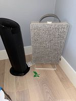 View of black Lasko tower heater and rectangular IKEA air purifier with light gray fabric front, both placed on wooden floor near wall corner.
