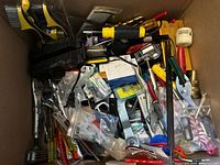 Top-down view into a box showing multiple hand tools including pliers, screwdrivers, a soldering iron, drill bits, and various small hardware items and packages scattered inside.