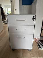 Front view of white metal filing cabinet showing three drawers and lock with keys inserted.