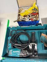 Makita rotary hammer tool with cord and side handle inside open hard plastic teal Makita case, drill bits visible. Additional container with mixed soldering and welding accessories behind.