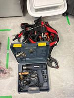 Blue Mastercraft heat tool in molded plastic case with cord and nozzle attachments on floor alongside red and black bag of clamps and tools.